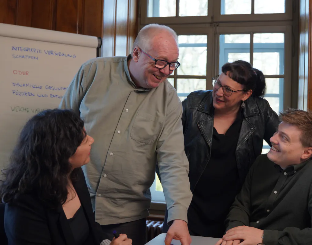 Vier Personen im Gespräch an einem Tisch, daneben ein Flipchart mit handschriftlich notierten Varianten zur integrierten Versorgung.